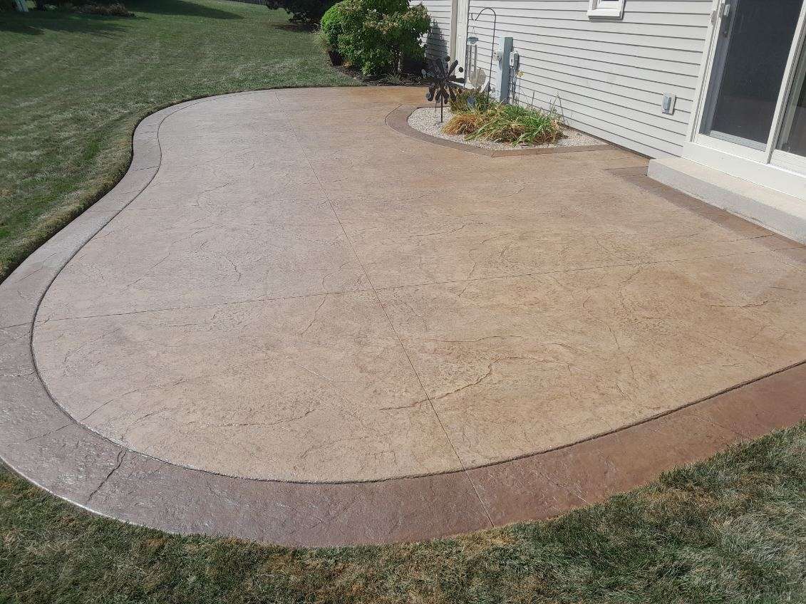 Curved concrete patio beside a house, bordered by grass and a small garden bed.