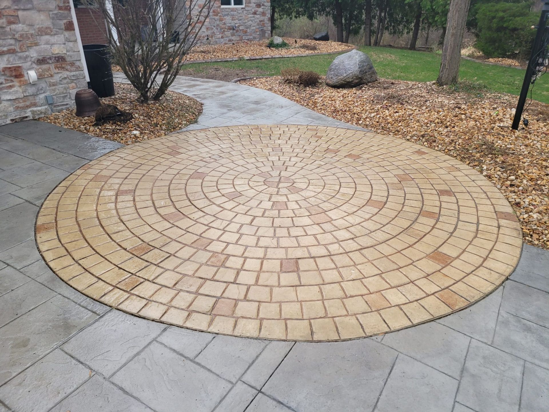 Circular brick patio in a landscaped yard beside a stone walkway and building.