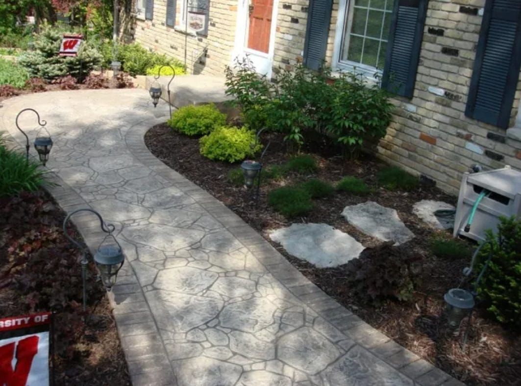 Stone walkway leading to a brick house with shrubs and garden beds in sunlight