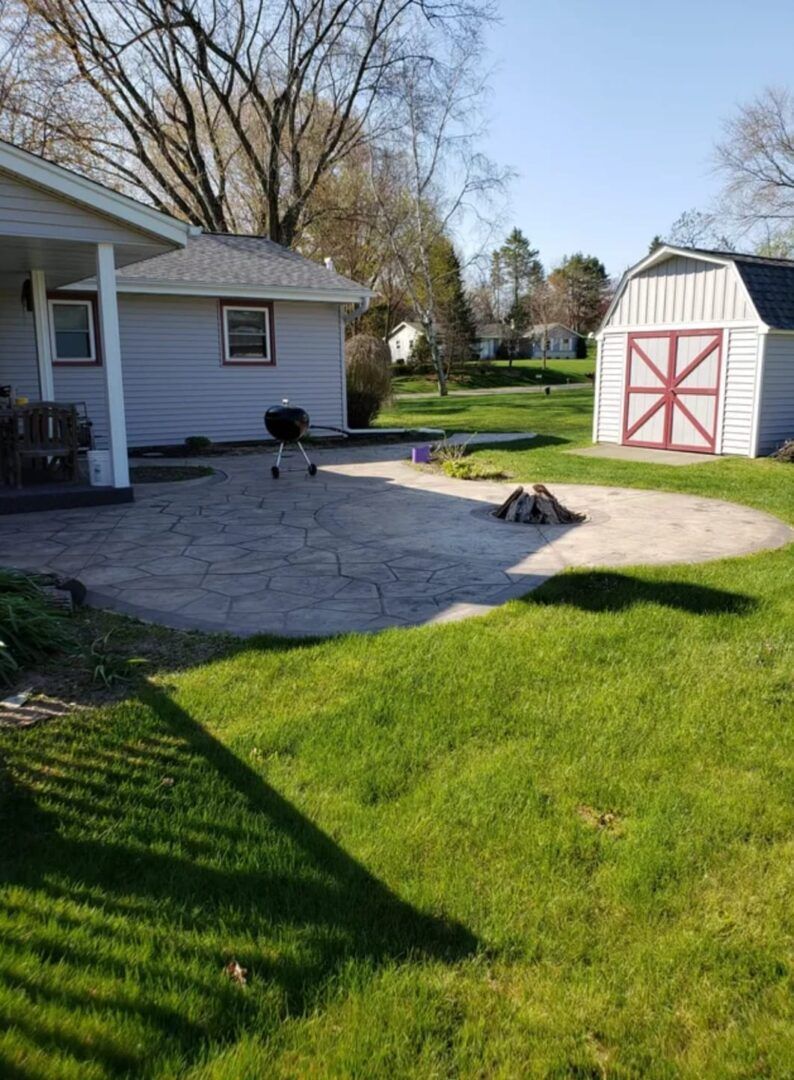 Backyard patio with house, lawn, and white shed with red trim in sunlight
