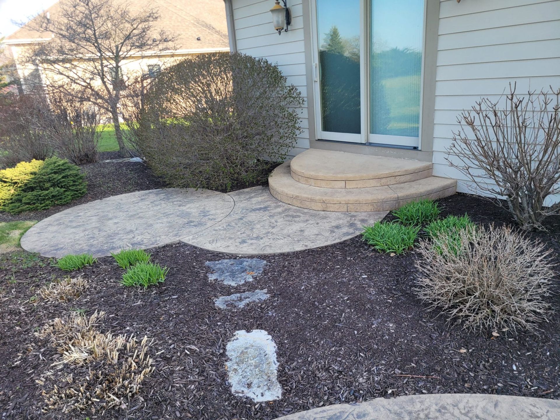 Front yard garden bed with mulch, shrubs, and stone walkway leading to a house patio door.