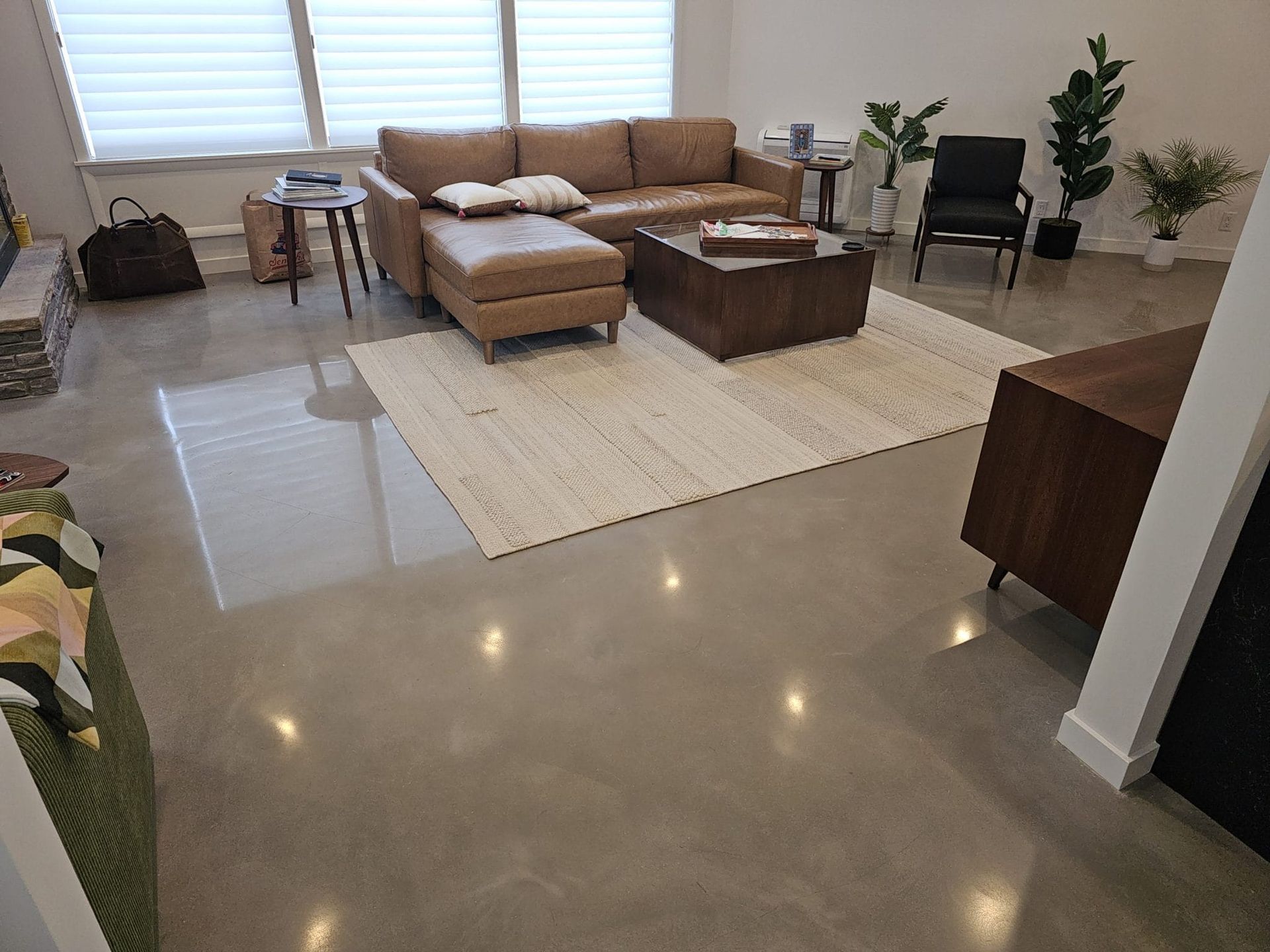 Spacious living room with tan sectional, ottoman, cream rug, glossy tile floor, and potted plants by windows.