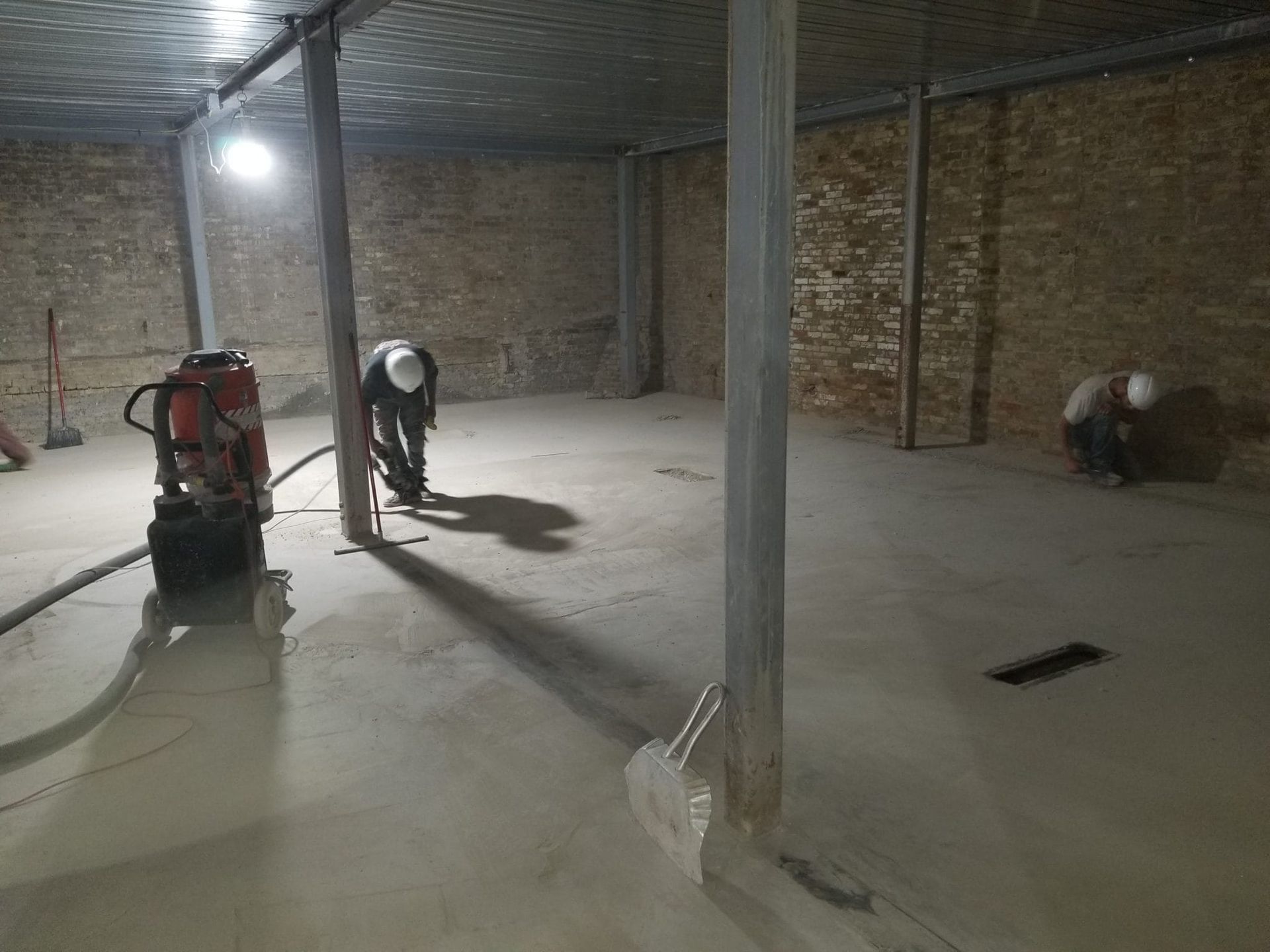 Workers sanding a concrete floor in a dim unfinished room with brick walls and support columns
