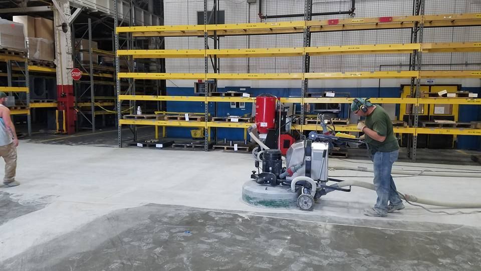 Worker operating a floor grinder in an industrial warehouse with yellow railings and concrete dust