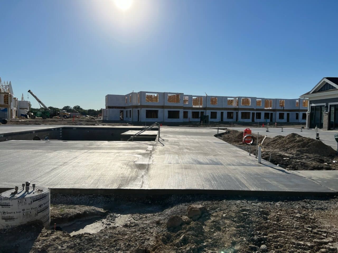 Sunny construction site with a concrete slab, dirt piles, and a row of townhouses in the background