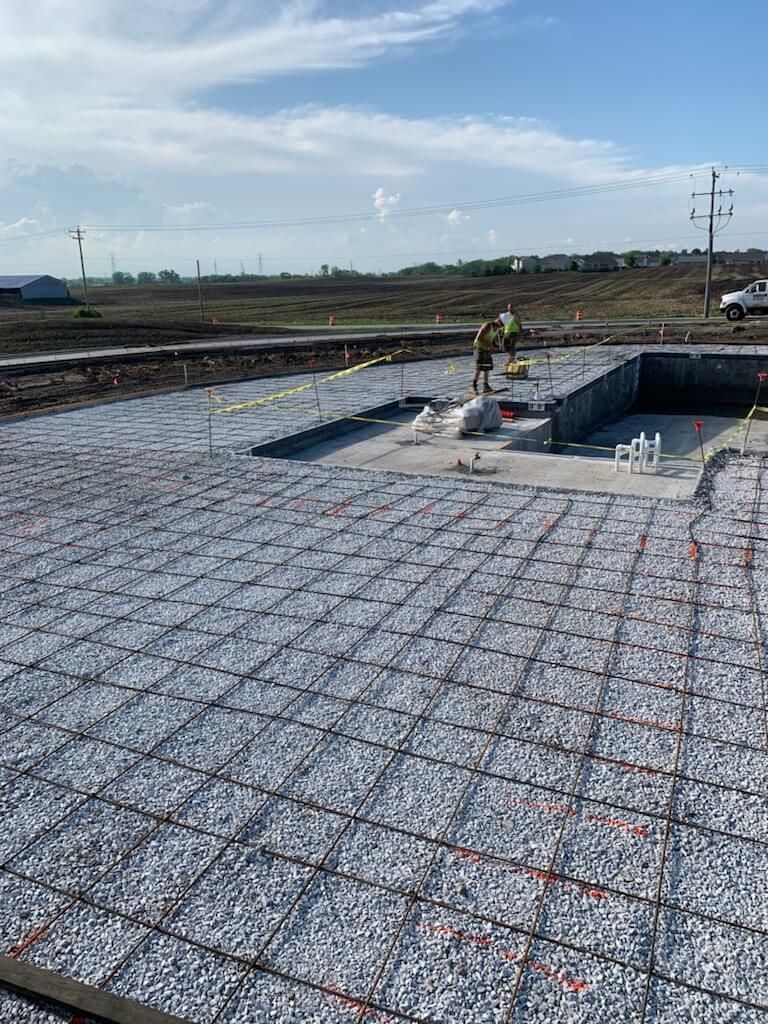 Construction site with rebar mesh over foundation slab and a worker near an excavated pit under a cloudy sky