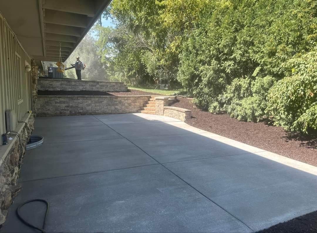 Concrete patio beside a stone retaining wall and tall green trees in a sunny yard