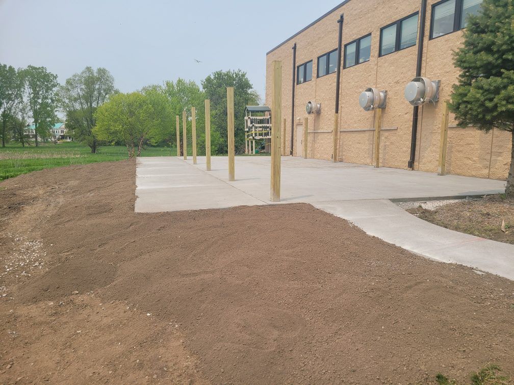 Brick building with new concrete walkway, dirt landscaping, and several wooden posts outside