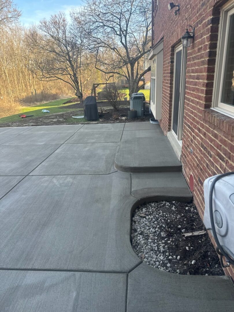 Concrete patio beside a brick house, with a small landscaped area and trees in the background.
