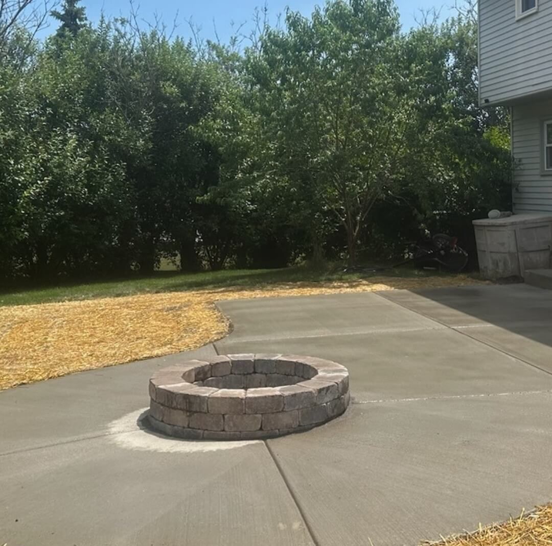 Concrete driveway with a circular stone fire pit in the center, beside trees and a house.