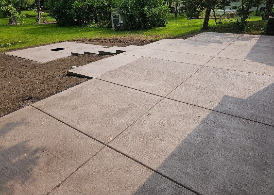 Concrete patio slabs beside a bare dirt section in a green yard, with trees in the background