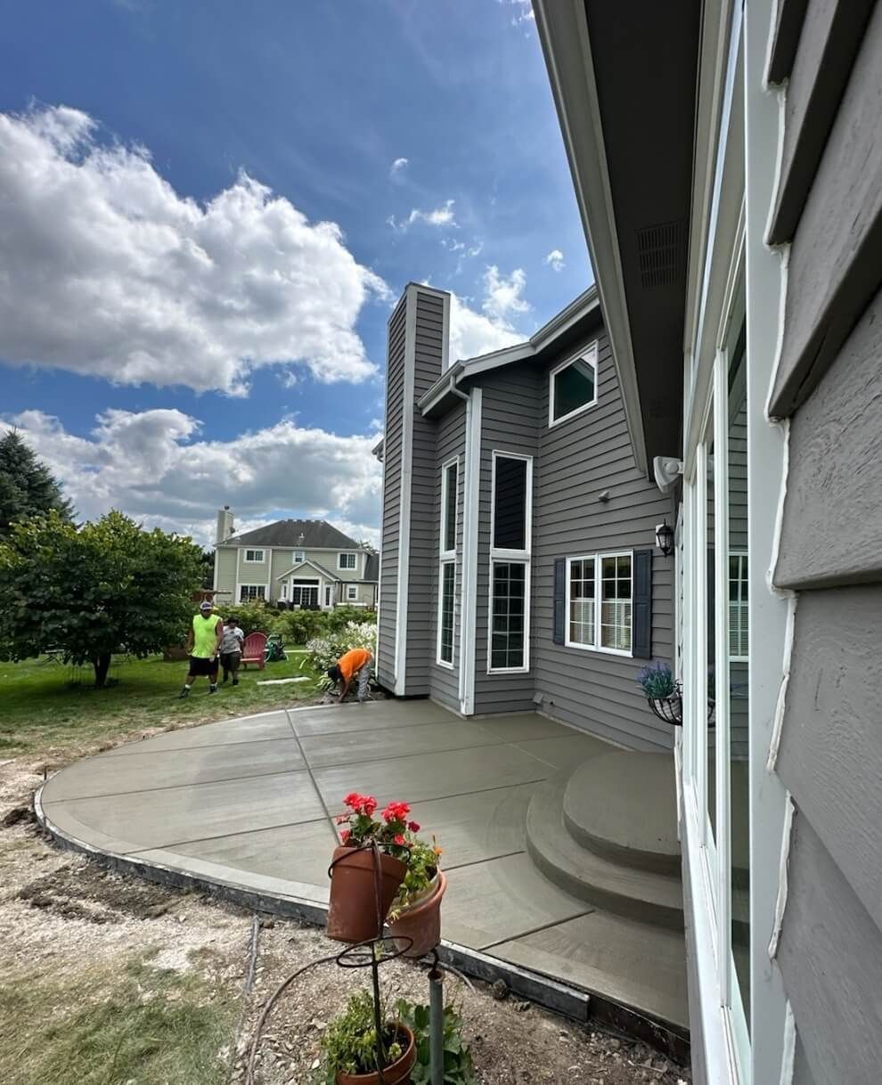 Gray house exterior with curved concrete patio, potted flowers, and people near a grassy yard under a blue sky