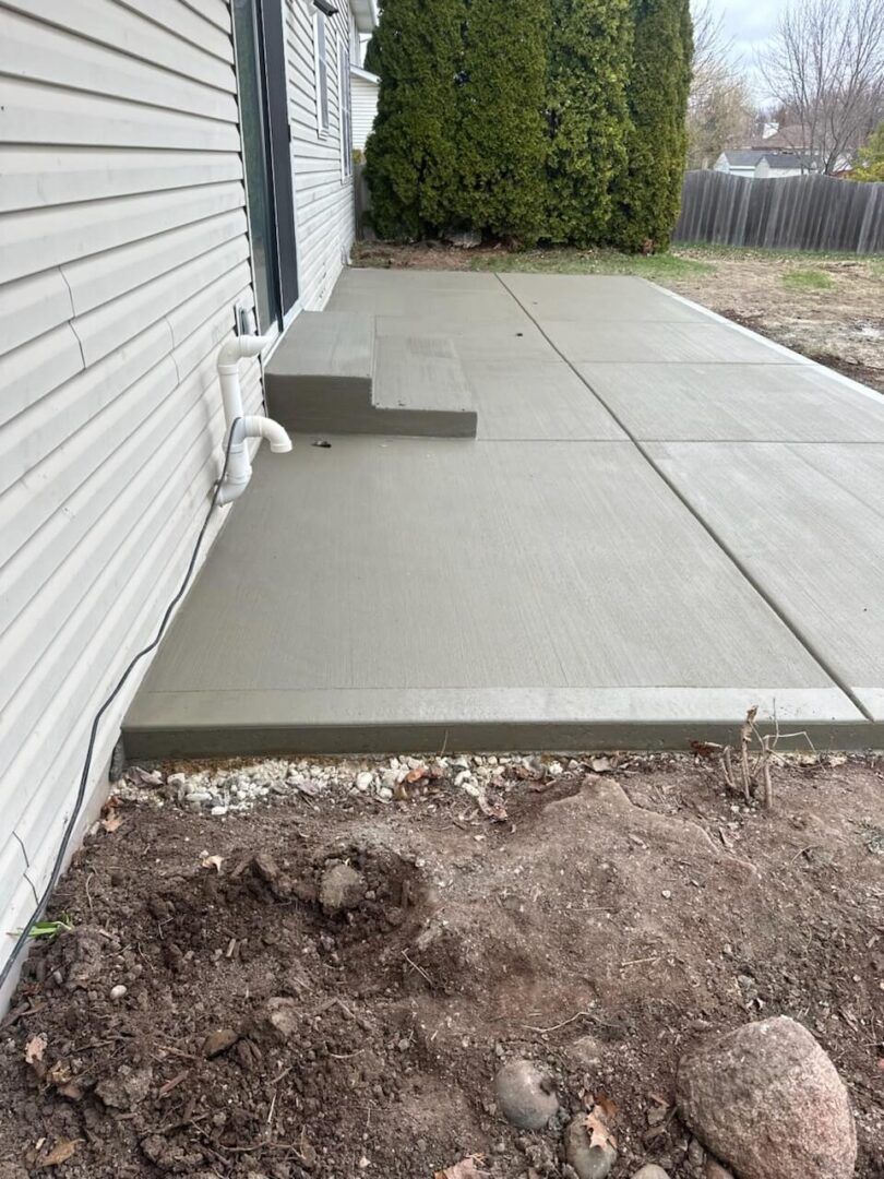 Concrete patio beside house with small step, hedge, and bare dirt in the foreground