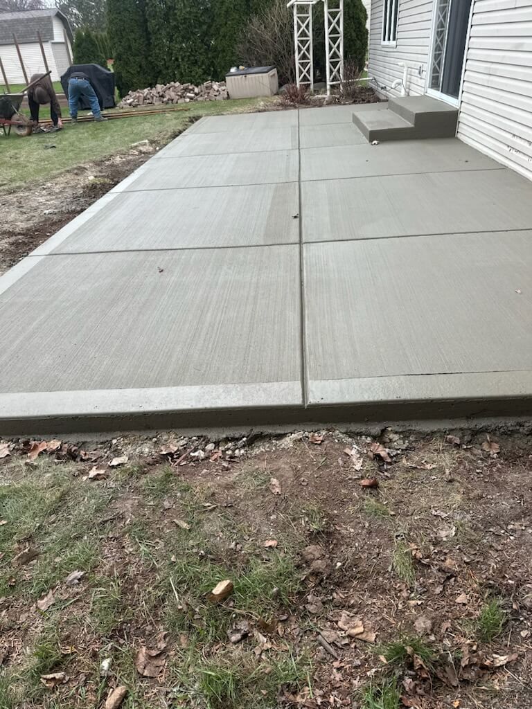 Concrete patio slabs beside a house, with a narrow garden border and yard in the foreground