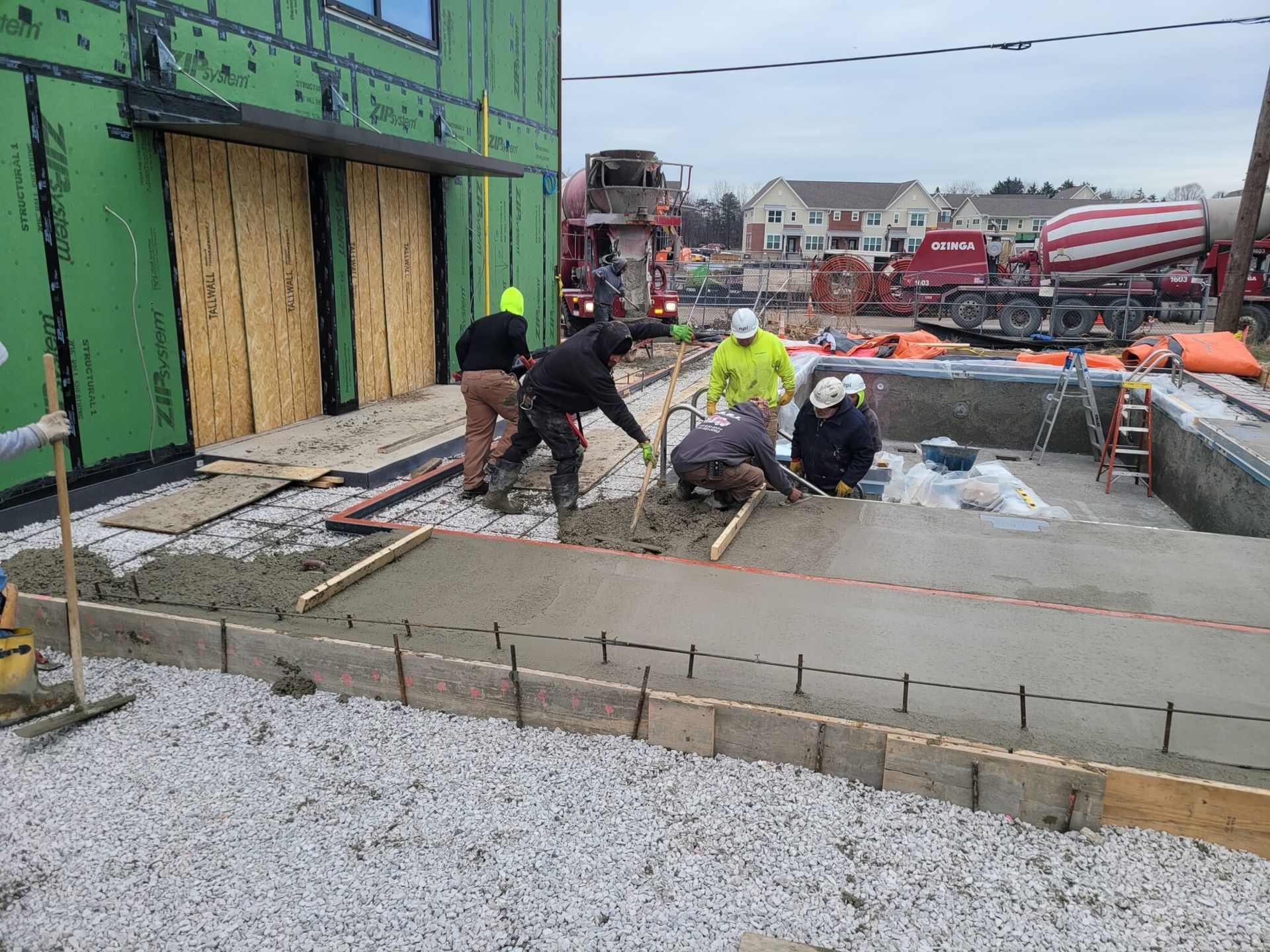 Construction workers pour concrete at a building site beside a green-framed wall.