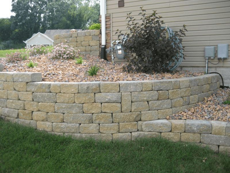 Tiered stone retaining wall with mulch bed, shrubs, and a house exterior in the background