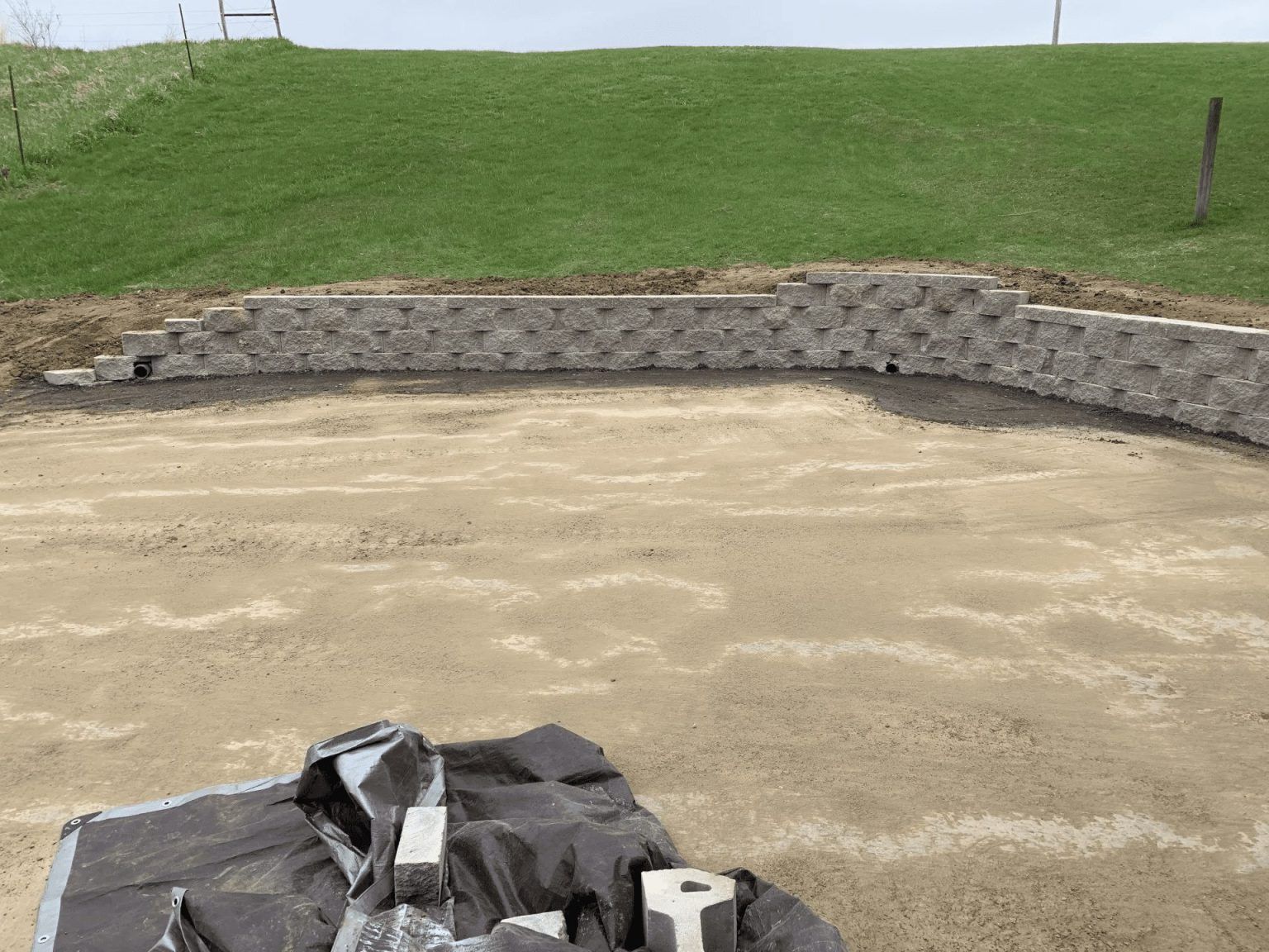 Sandy vacant lot with a stone retaining wall and grassy hill in the background