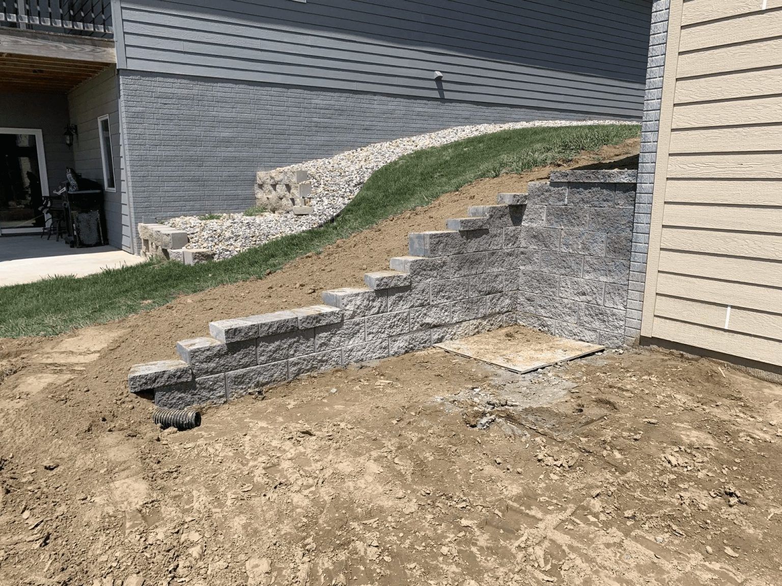 Stone retaining wall with stepped ledges along a grassy slope beside a beige house and dirt yard