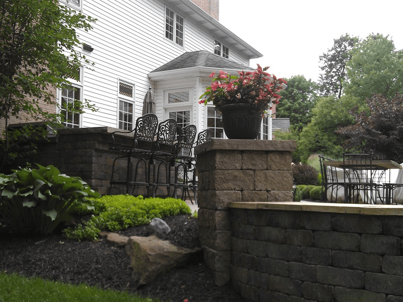 Stone patio with black wrought-iron chairs, flower planters, and a white house in the background