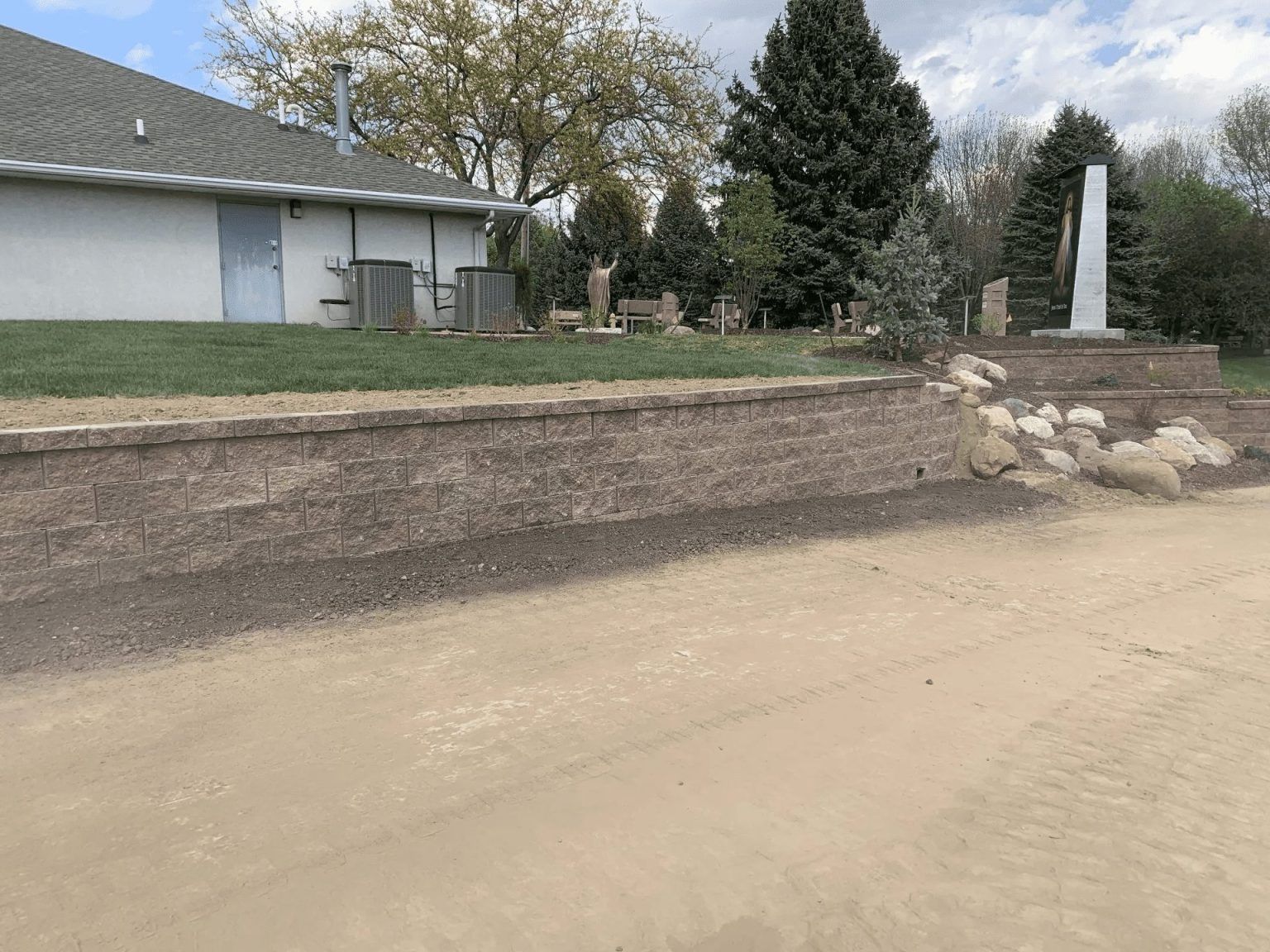 Eroded roadside embankment beside a house with a dirt road and stone steps under cloudy skies