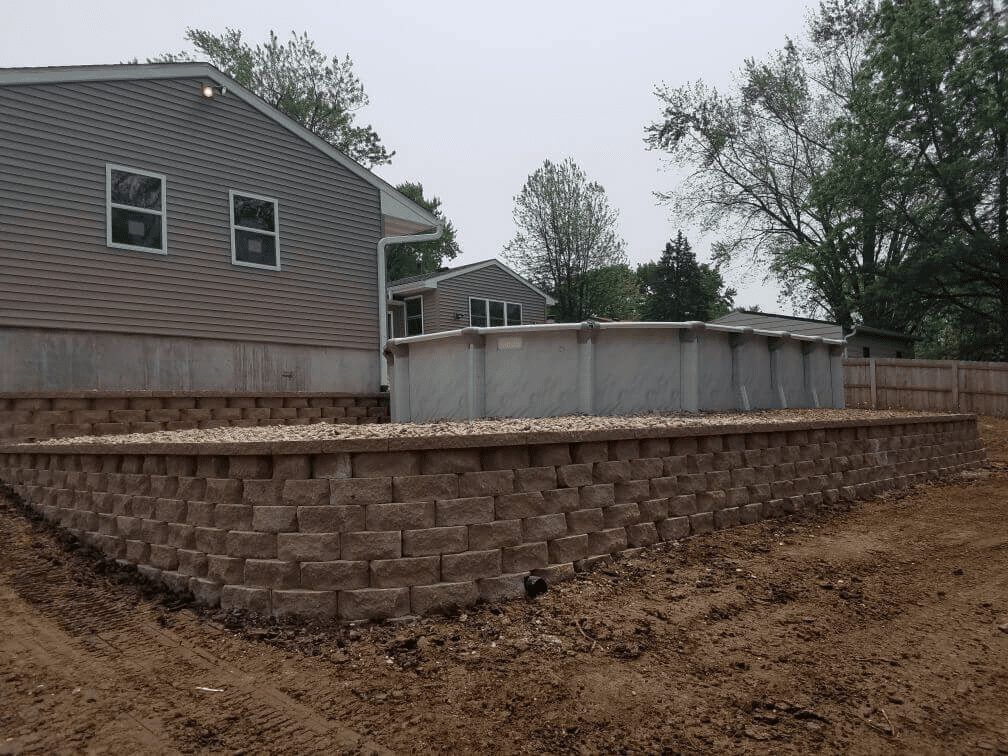 Retaining wall and patio beside a gray house under construction, with dirt yard and trees in back.