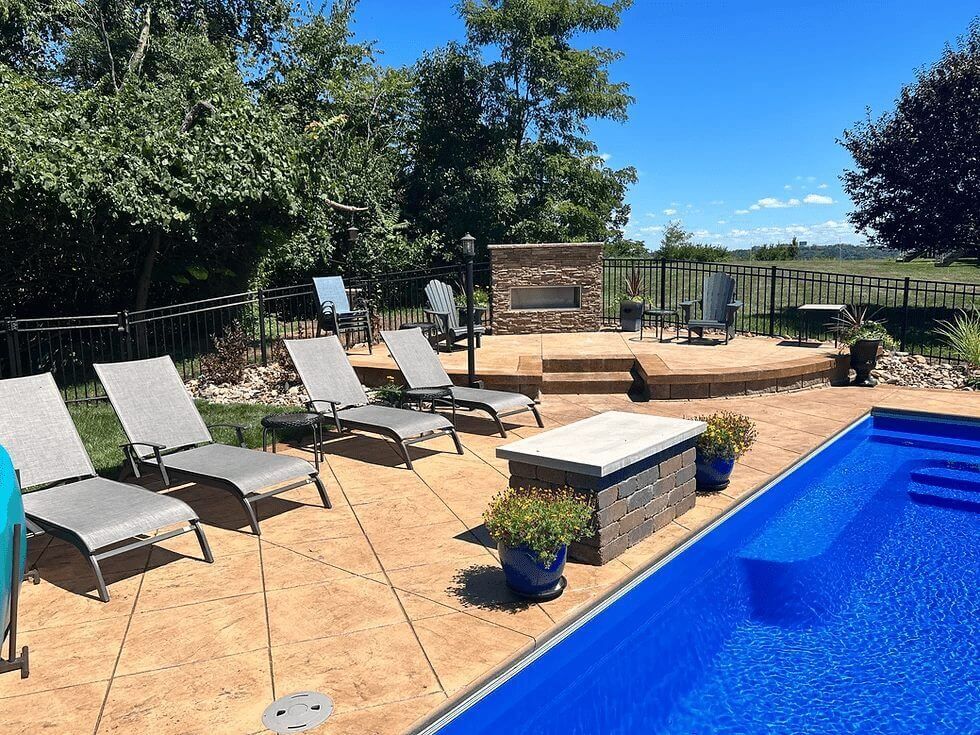 Backyard patio with lounge chairs, fire pit, and blue swimming pool under a sunny sky