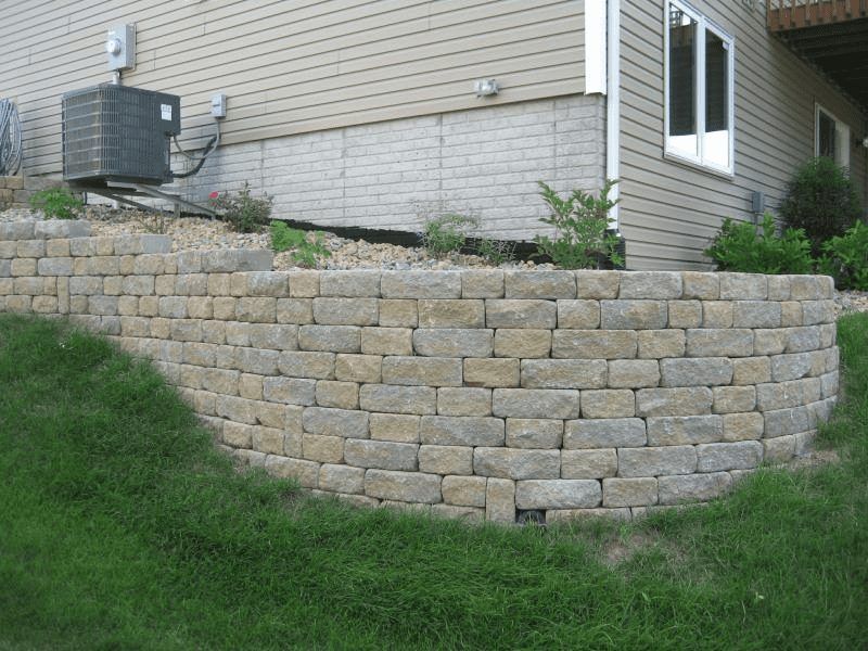 Stone retaining wall beside a house, with grass and shrubs on a sloped yard.