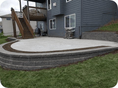 Curved concrete patio with stone retaining wall beside a gray house and wooden stairs in a backyard