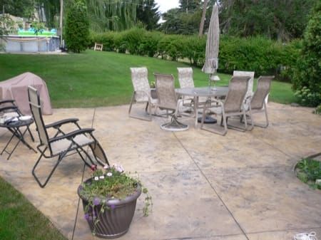 Patio with glass table, chairs, and lounge chairs on a concrete patio beside a green lawn and garden.