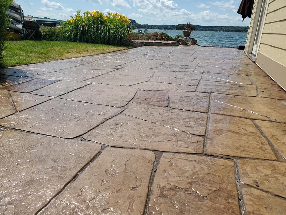 Stone patio beside a grassy yard and waterfront, with blue water and a dock in the distance.