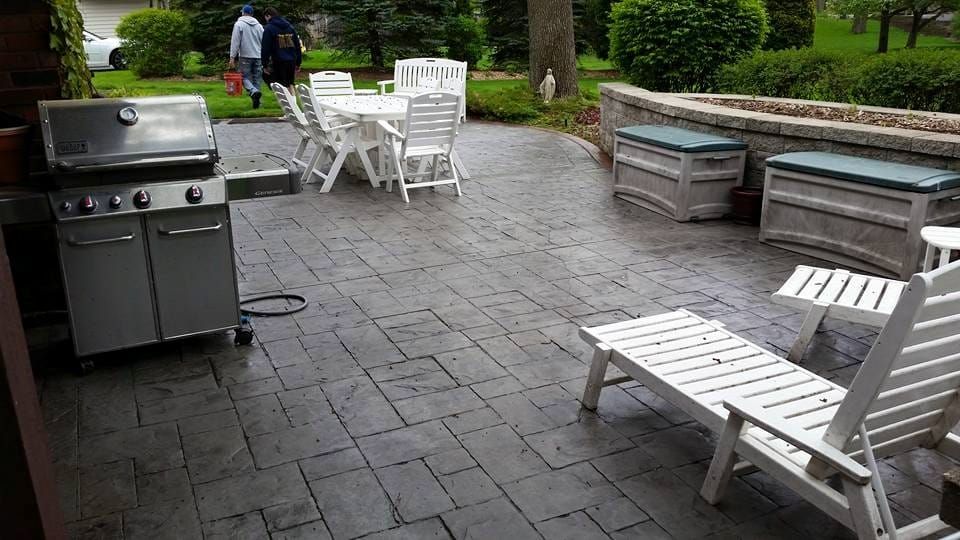 Patio with grill, white lounge chairs, tables, and benches on stone paving in a backyard