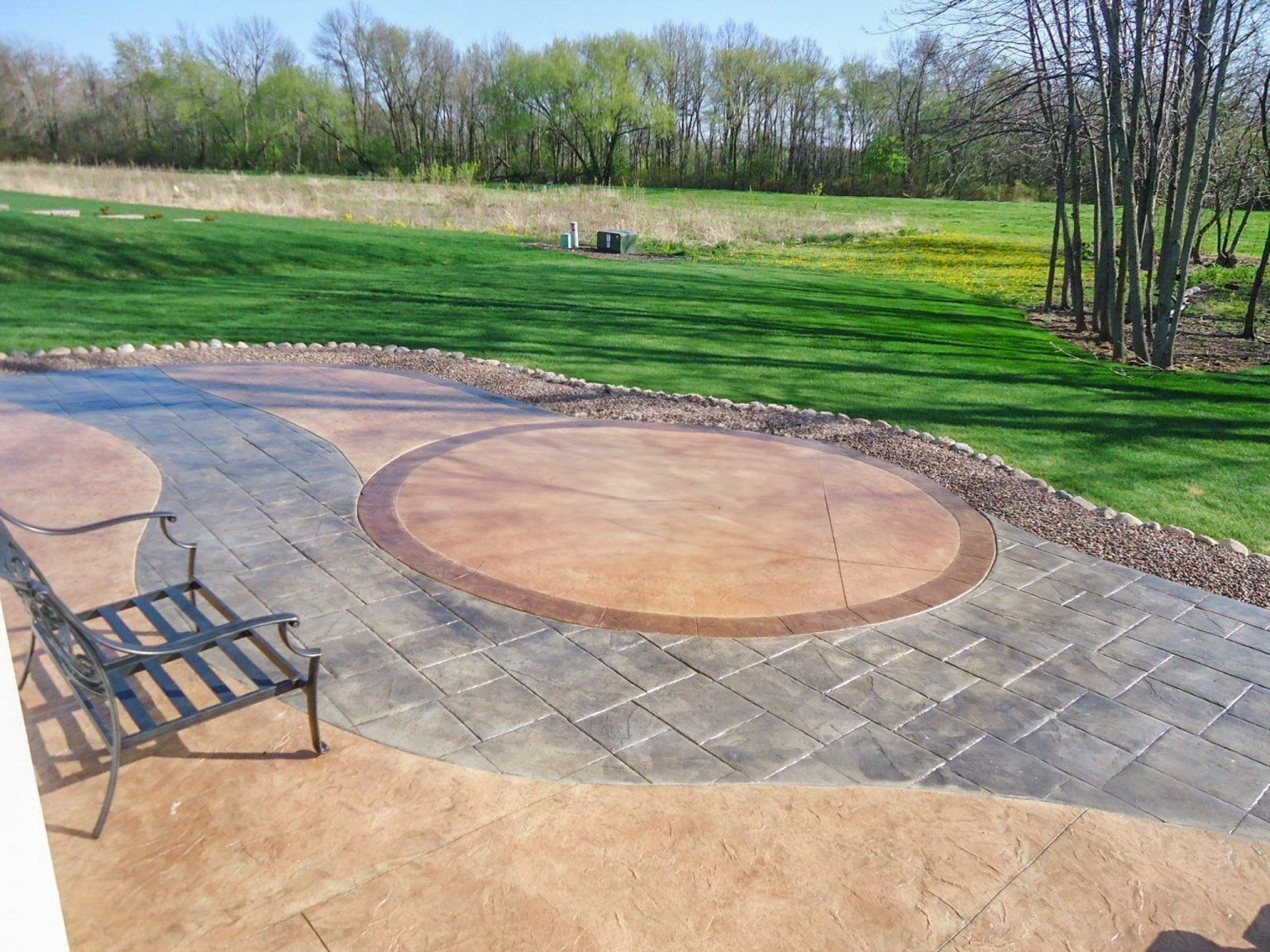 Patio with circular paver fire pit, bench, and green lawn in a park-like backyard