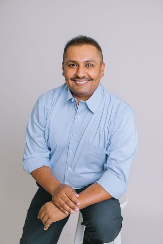 Smiling person seated against a light gray background, wearing a light blue button-up shirt and dark pants
