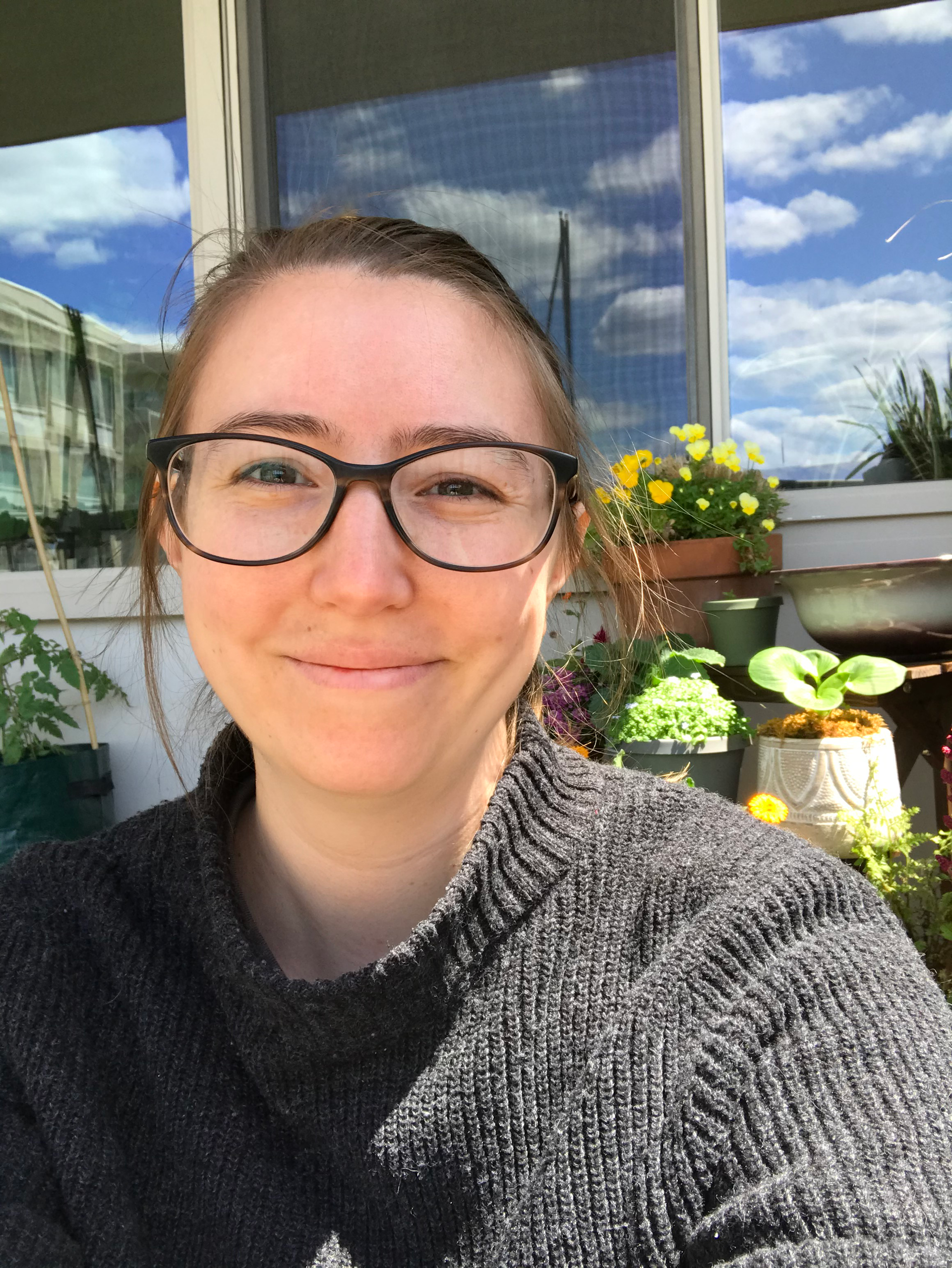 A woman wearing glasses and a sweater is smiling in front of a window.