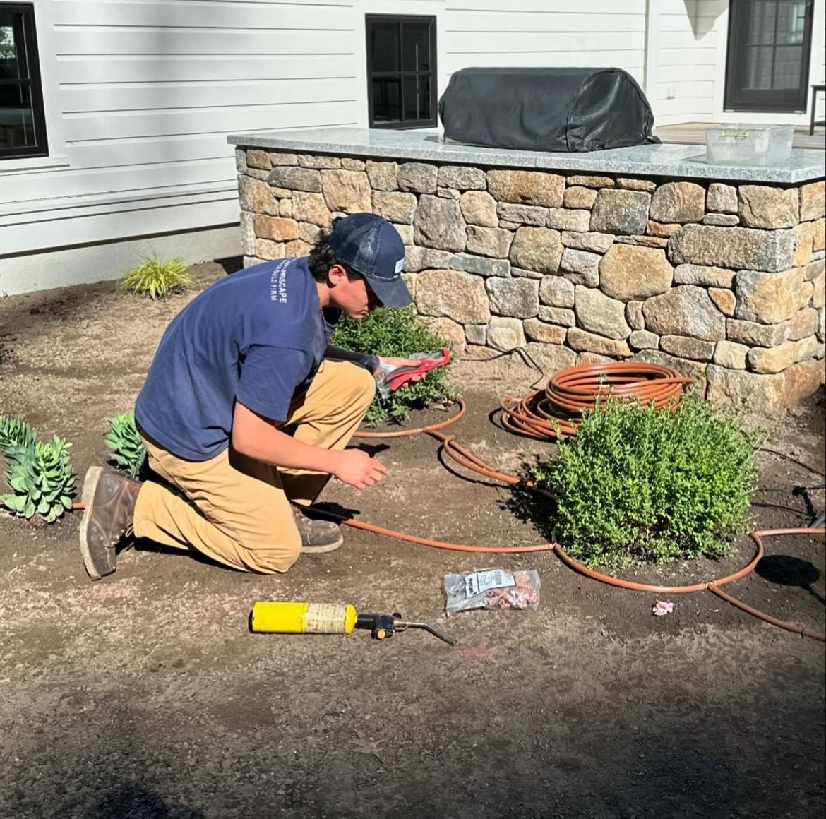 A man is kneeling down in the dirt in front of a stone wall.