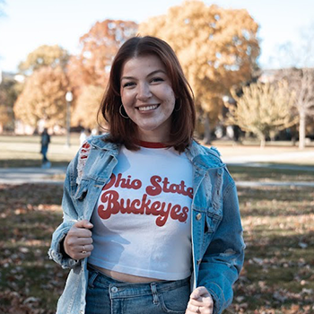 a woman wearing a ohio state buckeyes t-shirt and a denim jacket is standing in a park .