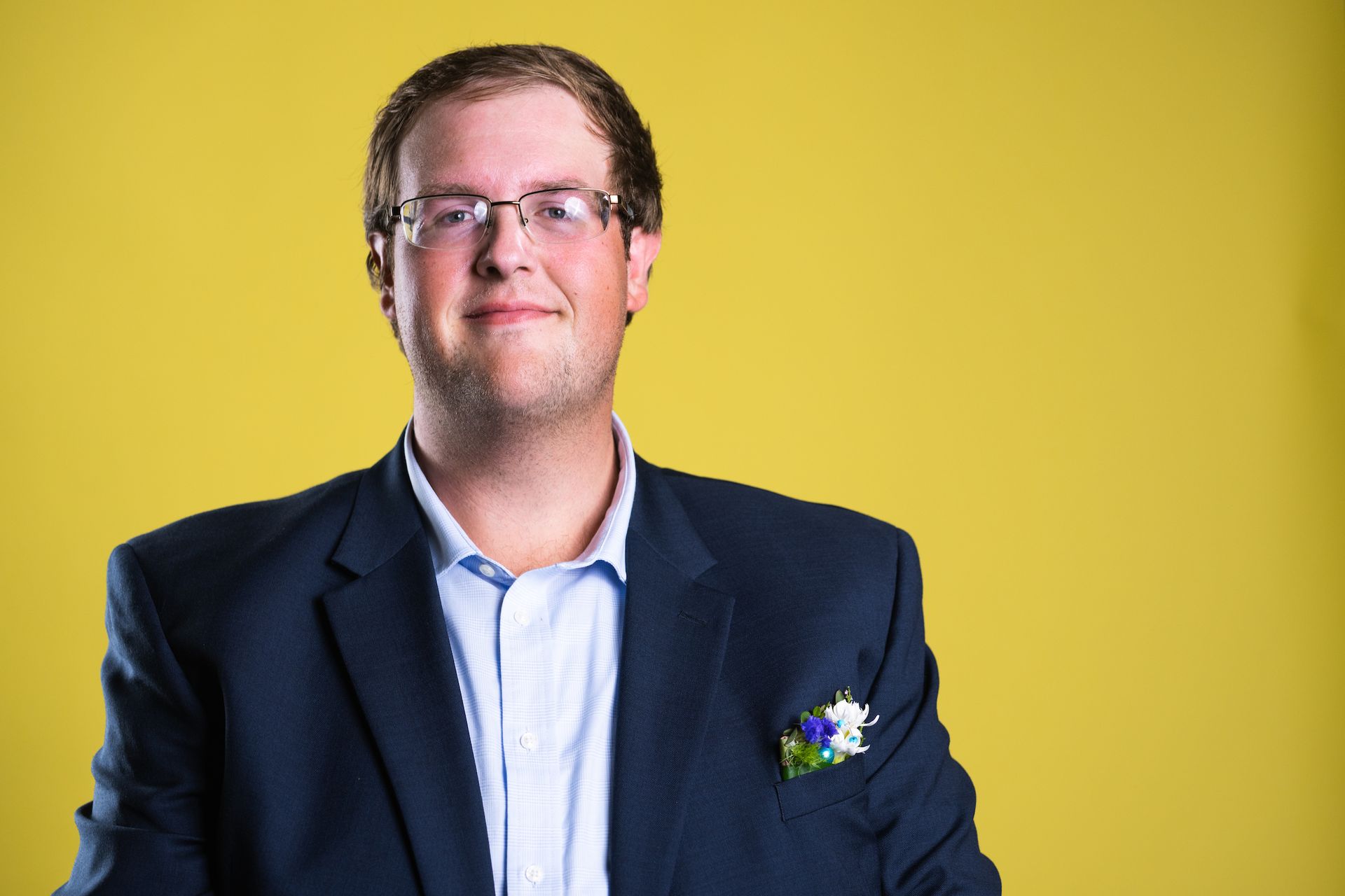 A man in a suit and glasses is standing in front of a yellow background.