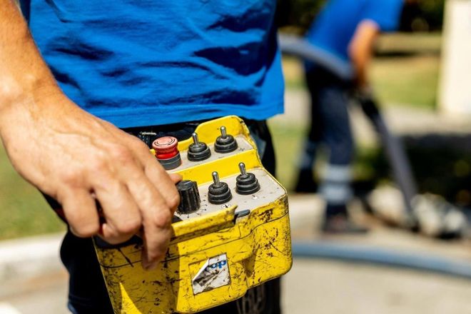 Persona che tiene in mano un pannello di controllo giallo con più interruttori. Un'altra persona lavora sullo sfondo con un tubo.