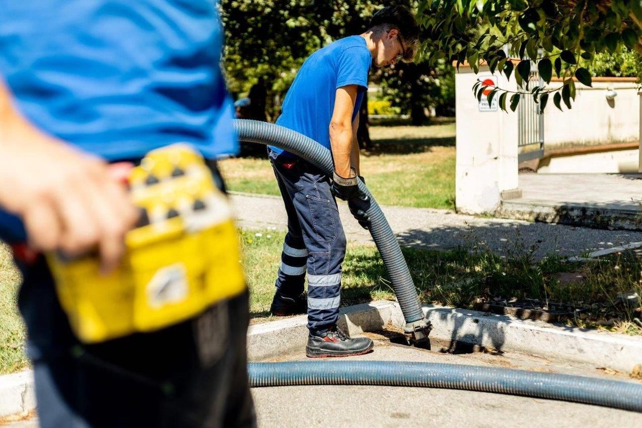 Due operai utilizzano un tubo flessibile per aspirapolvere vicino a un marciapiede; uno tiene in mano un pannello di controllo, l'altro è chino sul tubo.