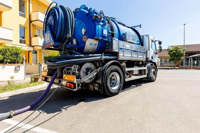 Un camion fognario blu e bianco parcheggiato in un'area aperta, con un tubo che arriva fino a terra.