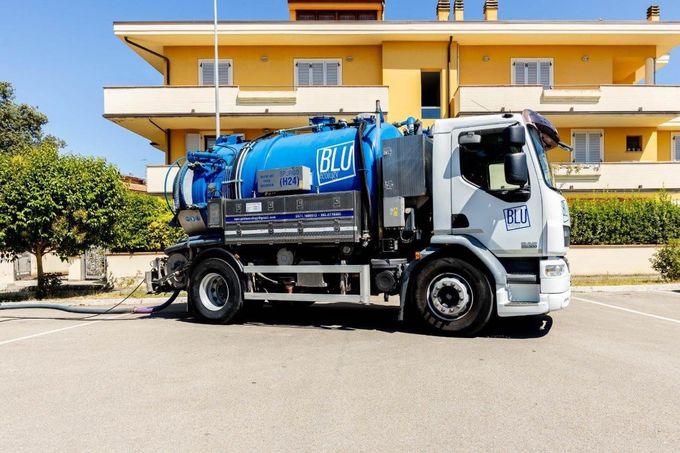 Un camion aspiratutto blu e bianco parcheggiato davanti a un edificio giallo in una giornata di sole.