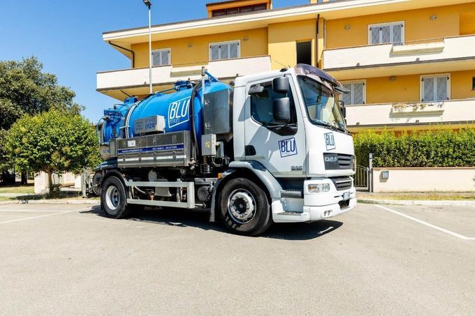 Un camion della spazzatura bianco e blu parcheggiato in un piazzale asfaltato di fronte a un edificio giallo in una giornata di sole.