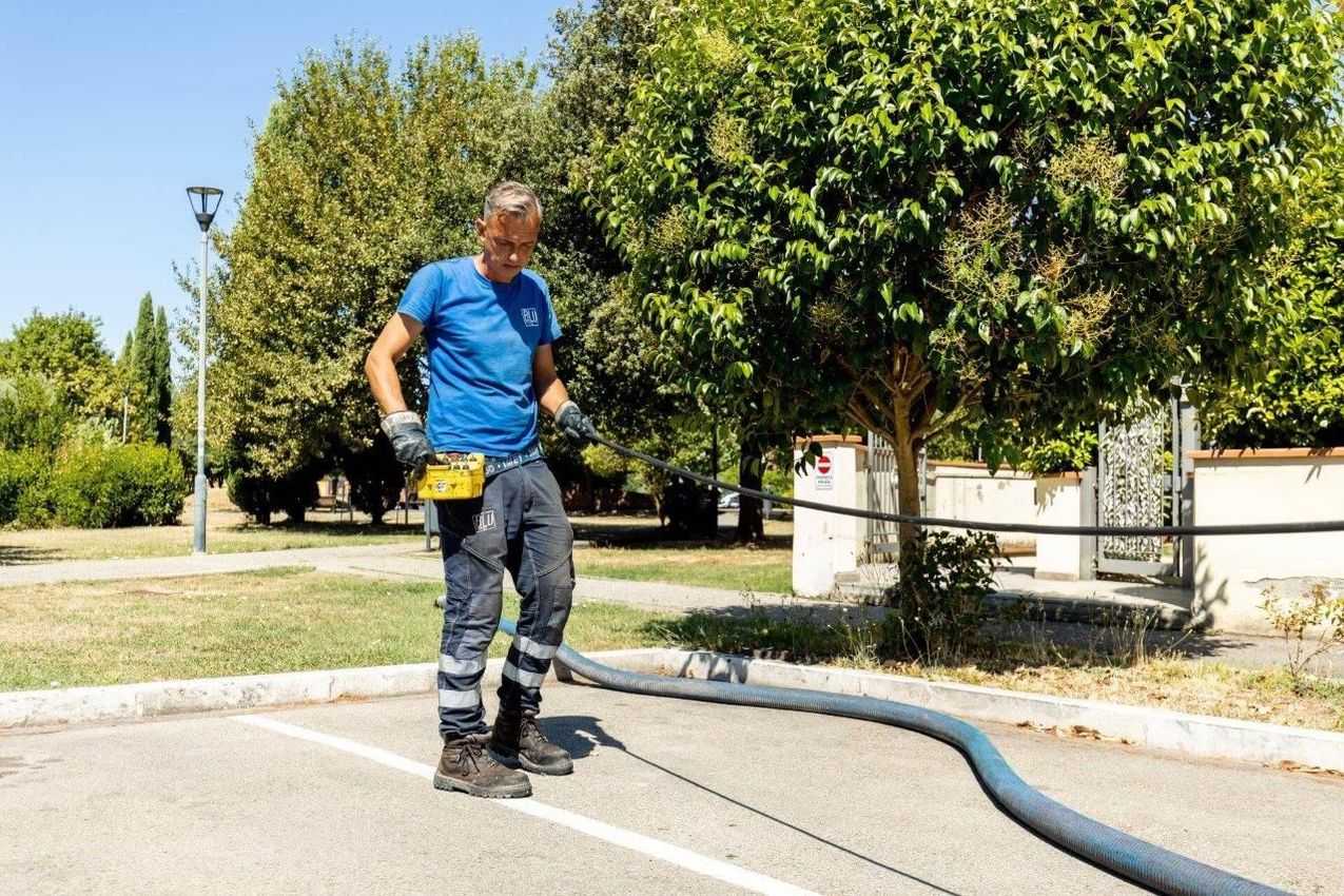 Un uomo in camicia blu e pantaloni da lavoro osserva un tubo su un'area pavimentata con alberi sullo sfondo.