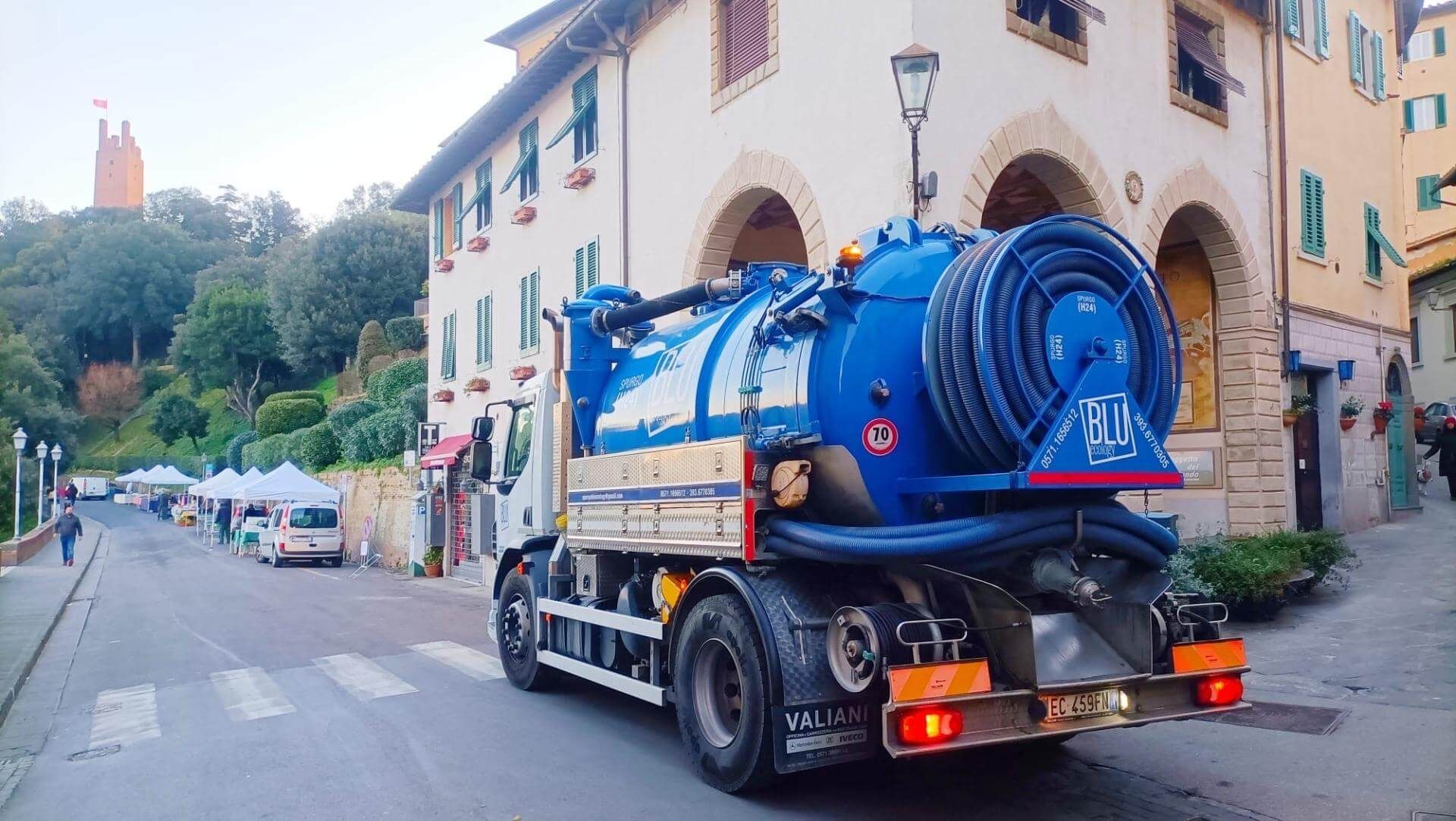 Camion blu per la pulizia della strada, in un contesto urbano. Edifici e alberi sullo sfondo.