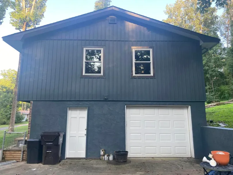 Two-story building with a garage. Dark blue siding, white garage door, two windows, and white door. Trees in the background.