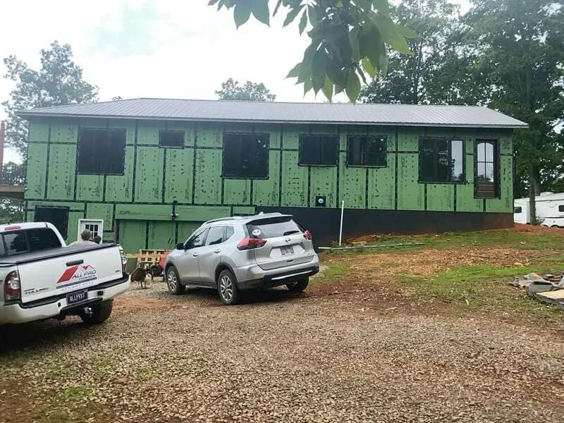 A house under construction with green insulation, dark windows, and a silver car in front.