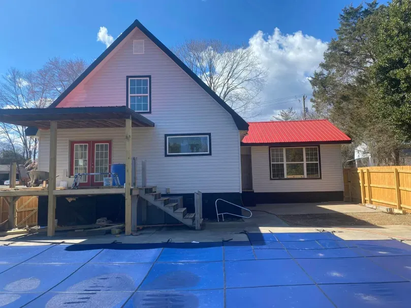 White house with black roof, red door, and red roofed addition. Blue tarp in foreground.
