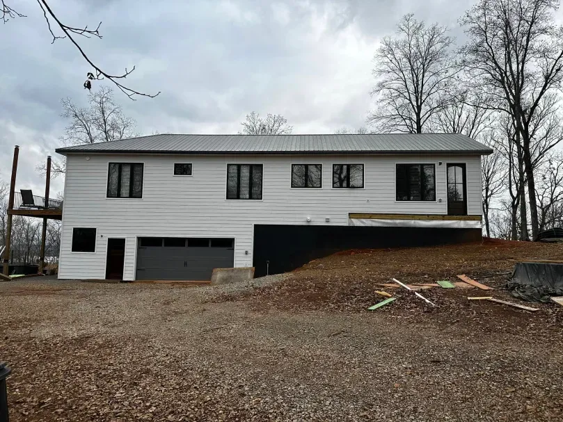 White two-story house with black windows and a metal roof on a hill with a gravel driveway.