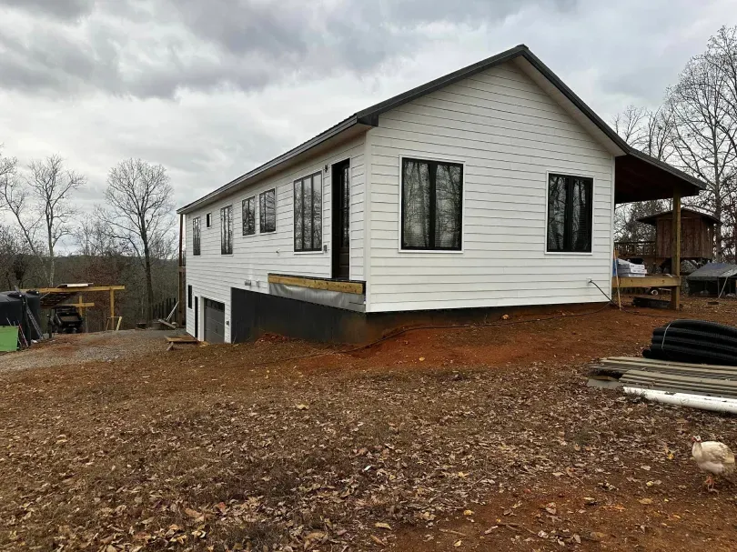 White house with black window frames, porch, and exposed foundation on a brown, sloped lot under an overcast sky.