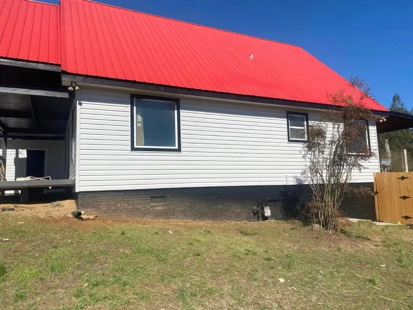 Side view of a house with white siding and a red roof, a small bush and window.